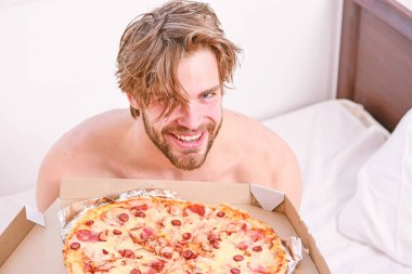 Adam pizza kutusu sit yatak yatak odası ya da otel odasında tutar. Evde yatakta lezzetli bir pizza yemek parlak bir apartman dairesinde öğrencidir. Gömleksiz seksi adam yatakta pizza ile görüntü kırpılmış. Yatakta yemek.