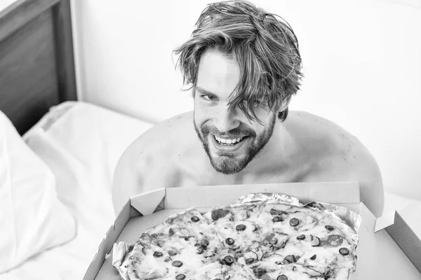 Guy holds pizza box sit bed in bedroom or hotel room. Student is at home on the bed in a bright apartment eating a tasty pizza. Cropped image of shirtless sexy man with pizza on bed. Dinner in bed.