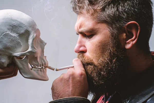 Habit to smoke tobacco bring harm to your body. Smoking cause health damage and death. Man smoking cigarette near human skull symbol death. Nicotine destroy health. Harmful habits. Smoking is harmful