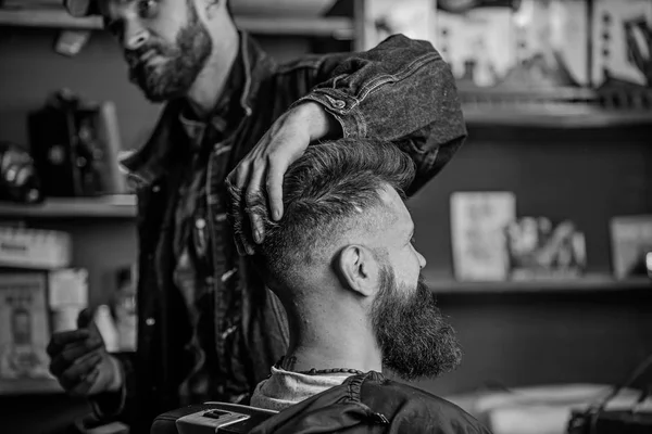 Hipster client with fresh haircut or hairstyle. Barber styling hair of bearded client with wax by hands. Man with beard and mustache in hairdressers chair, shelves on background. Barbershop concept — Stock Photo, Image