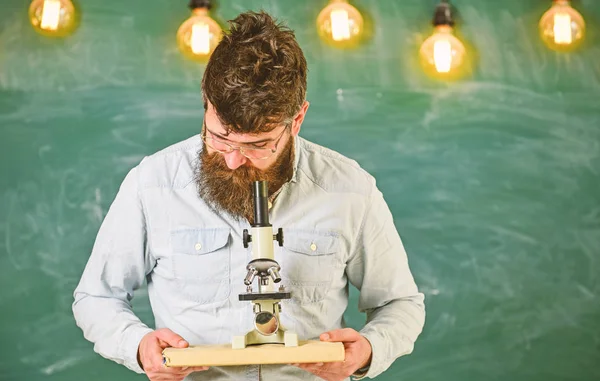 Hombre con barba y bigote en la cara ocupada. Científico sostiene libro y microscopio, pizarra en el fondo. Concepto de investigación científica. Profesor en anteojos sostiene libro y microscopio — Foto de Stock