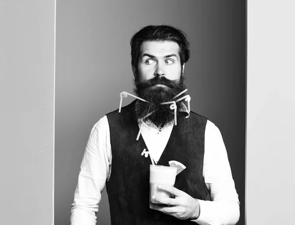 handsome bearded man with long beard and mustache has stylish hair on surprised face holding glass of alcoholic beverage in vintage suede leather waistcoat on colorful studio background, copy spac