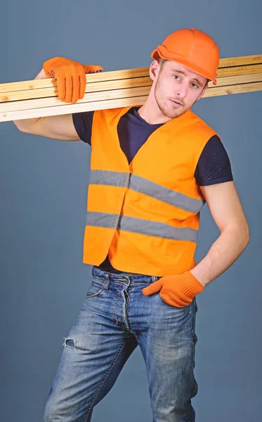 Carpintero, carpintero, obrero, constructor de cara segura lleva vigas de madera en el hombro. Hombre con casco, sombrero duro y guantes de protección sostiene la viga de madera, fondo gris. Concepto de trabajador resistente — Foto de Stock