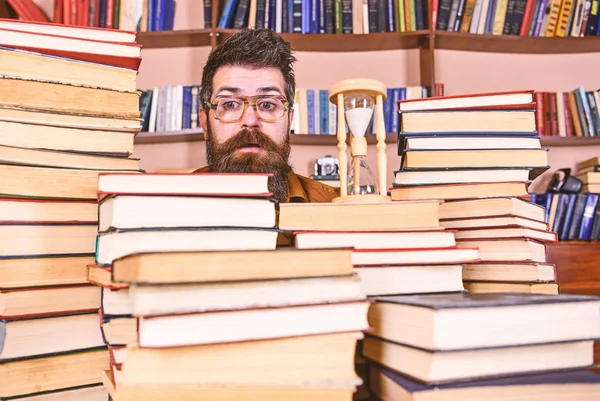 Conceito de fluxo de tempo. Professor ou estudante com barba estudando na biblioteca. Meu, cientista de óculos olha para a ampulheta. Homem no rosto surpreso senta-se perto de ampulheta enquanto estuda, estantes de livros no fundo — Fotografia de Stock