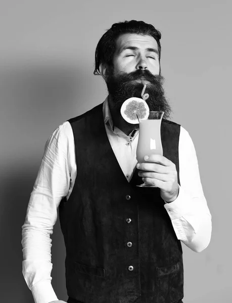 handsome bearded man with long beard and mustache has stylish hair on satisfied face holding glass of alcoholic cocktail in vintage suede leather wa