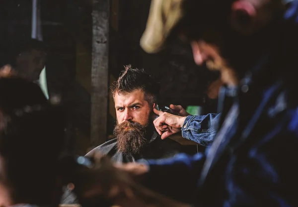 Barber with hair clipper works on hairstyle for man with beard, barbershop background. Barber styling hair of brutal bearded client with clipper. Haircut concept. Hipster client getting haircut