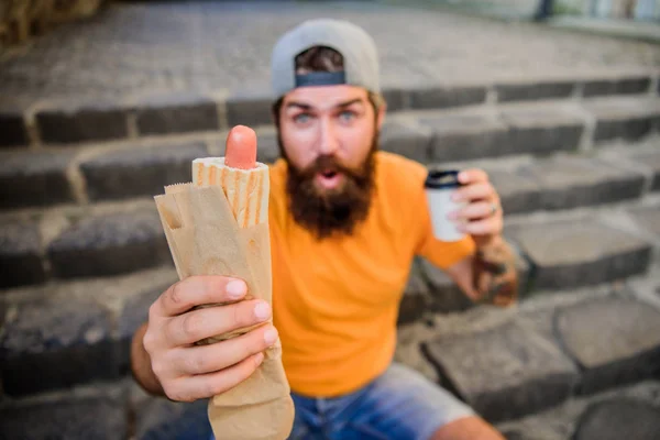 Fast food for fast times. Caucasian guy enjoying street food cuisine. Bearded man eating unhealthy junk food with takeaway drink. Hipster snacking with fast food during meal break
