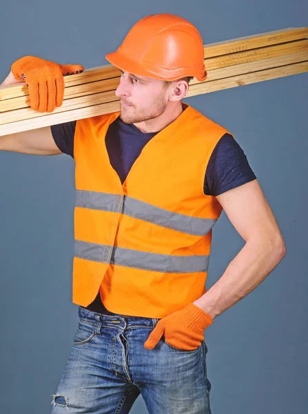 Σκληραγωγημένο εργάτης έννοια. Ξυλουργός, εργάτης, ξυλουργός, οικοδόμος στο πολυάσχολο πρόσωπο ασκεί ξύλινα δοκάρια στον ώμο. Ο άνθρωπος στο κράνος, προστατευτικά γάντια κατέχει ξύλινο δοκάρι, σκληρό καπέλο και γκρι φόντο — Φωτογραφία Αρχείου