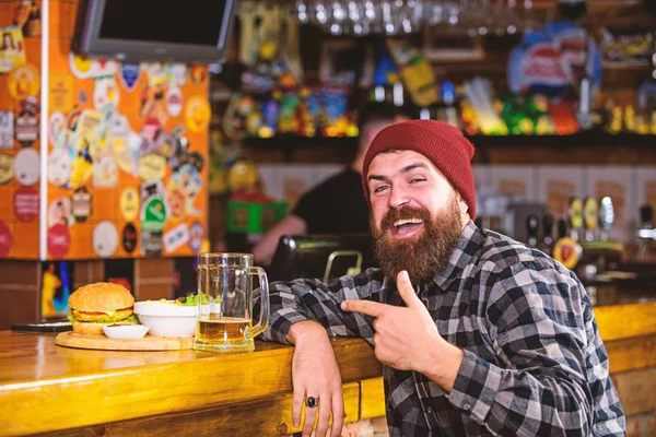 Pub is relaxing place to have drink and relax. Man with beard drink beer eat burger menu. Enjoy meal in pub. High calorie snack. Brutal hipster bearded man sit at bar counter. Hipster relaxing at pub