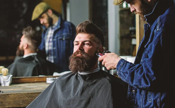 Hipster z brodą pokryte cape przycinania przez zawodowych fryzjera w stylowe fryzjera. Koncepcja oporządzanie konia. Fryzjer zajęty strzyżenie broda hipster klienta, lusterko refleksji na tle — Zdjęcie stockowe