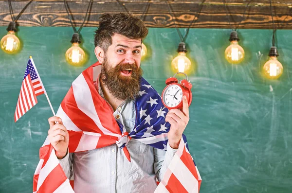 Vlag van de Verenigde Staten en klok, schoolbord houdt man met baard op vrolijk gezicht op achtergrond, kopie ruimte. Amerikaanse lerares met Amerikaanse vlaggen houdt wekker. Amerikaanse onderwijssysteem concept — Stockfoto