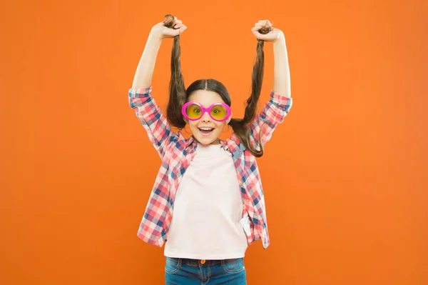 Eftersom dess ditt hår. Bedårande flicka som innehar ponytails hår på Orange bakgrund. Liten söt tjej med långt brunett hår bär fancy glasögon. Liten hårmodell med skönhetslook — Stockfoto