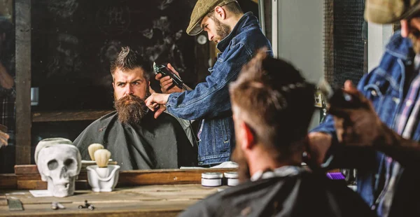 Concepto de corte de pelo. Peluquero con cortapelos trabaja en peinado para hombre con barba, fondo barbería. Hipster cliente cortarse el pelo. Peluquero peinado cabello de brutal cliente barbudo con clipper — Foto de Stock