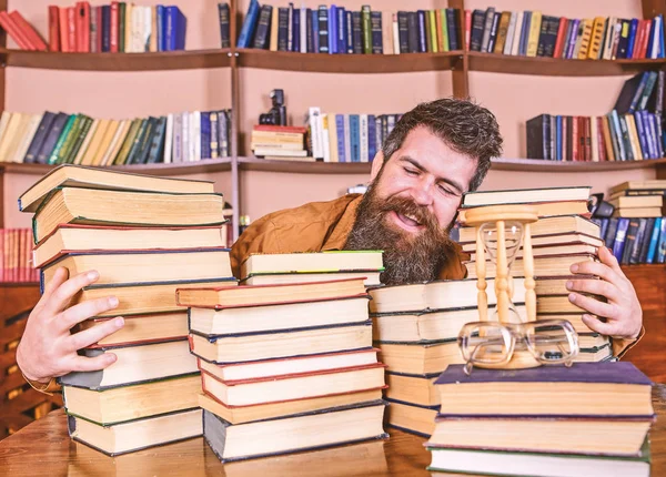 Βιβλιοθηκονόμος έννοια. Ο άνθρωπος στο χαρούμενο πρόσωπο μεταξύ σωρούς από βιβλία, ενώ σπούδαζε στη βιβλιοθήκη, ράφια σε φόντο. Δάσκαλος ή μαθητής με γενειάδα που κάθεται στο τραπέζι με τα βιβλία, defocused — Φωτογραφία Αρχείου