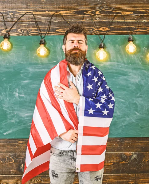 Концепция патриотического образования. Американский учитель, покрытый американским флагом. Учитель учит любить родину, США. Человек с бородой и усами на серьезном лице с флагом США, доска на заднем плане — стоковое фото