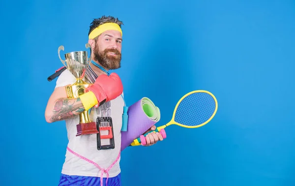 Man bearded athlete hold sport equipment jump rope fitness mat boxing glove expander racket and golden goblet. Choose sport you like. Sport shop assortment. Sport concept. Get body ready for summer