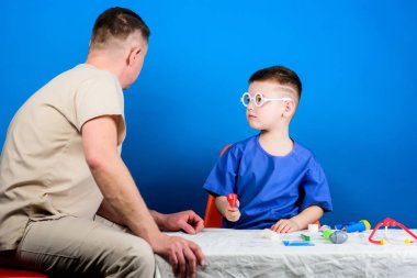 Tatlı çocuk ve babası doktor. Hastane çalışanı. Sağlık hizmetleri. Tıp konsepti. Küçük doktor, masada tıbbi aletlerle oturuyor. Hastalık tedavisi. Baba ve oğul tıp hanedanlığı. Tıbbi muayene