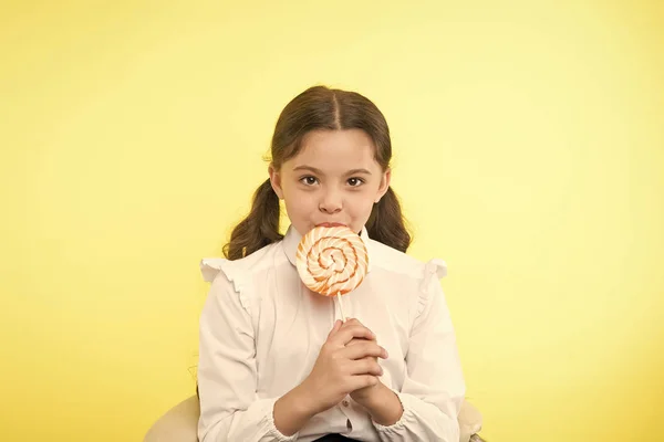 Unterernährung häufiges Problem. Mädchen Schüler Schuluniform mag Süßigkeiten Lollipop Süßigkeiten gelben Hintergrund. Mädchen niedlichen Kind Pferdeschwänze Frisur essen süßen Lutscher. Süßigkeiten in angemessenen Portionen ok — Stockfoto