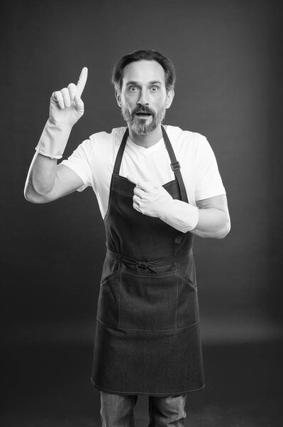 Просто посмотри туда. Пожилой человек, показывающий пальцем в жёлтых перчатках. Зрелый домашний работник представляет кое-что. Старший мужчина в фартуке и резиновых перчатках. Предоставление услуг по дому
