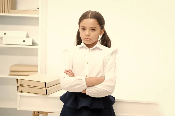 Little but serious. Child girl wears school uniform standing with crossed arms on chest. Schoolgirl smart child looks serious white interior background. Girl serious about knowledge and education