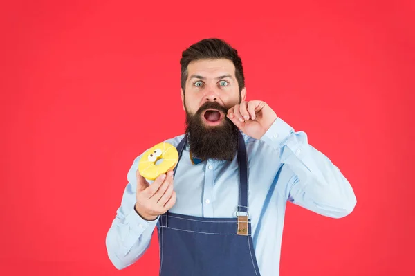 Cafe and bakery concept. Sweet donut from baker. Man bearded baker in cooking apron hold cute dessert. Forget about diet. Cheat meal. Hipster bearded baker hold glazed donut on red background