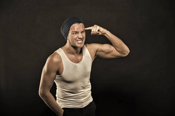 Man with muscular arms pointing at head with index finger. Masculinity concept. Macho on aggressive face with strong muscles look brutal, black background. Man in hat and sloven sleeveless undershirt