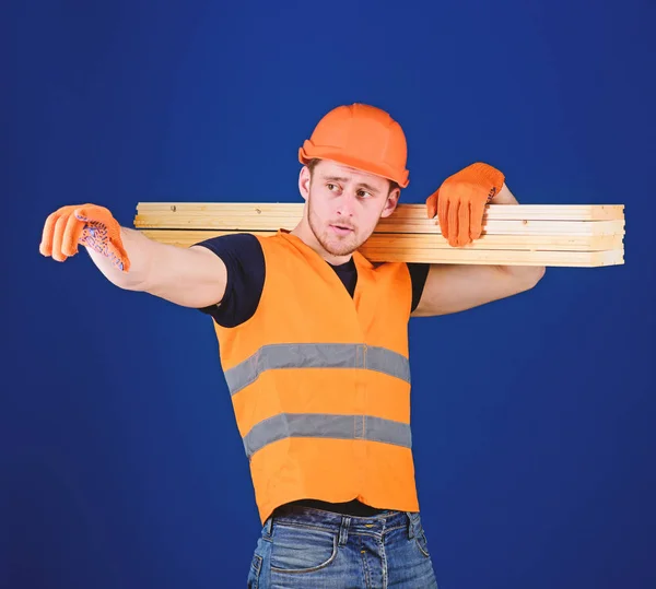 Man in helmet, hard hat and protective gloves pointing direction, blue background. Carpenter, woodworker, strong builder on thoughtful face carries wooden beam on shoulder. Wooden materials concept — Stock Photo, Image