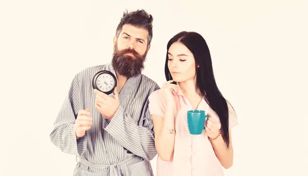 Chica con taza de café, el hombre tiene el reloj en la mano. Concepto matutino perfecto. Pareja, la familia se despertó a tiempo. Pareja enamorada, familia joven en pijama, albornoz aislado sobre fondo blanco — Foto de Stock