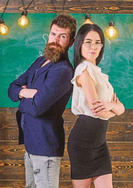 Man with beard and teacher in eyeglasses stand back to back, chalkboard on background. School staff concept. Lady and hipster working together in school. Teacher and schoolmaster look confident