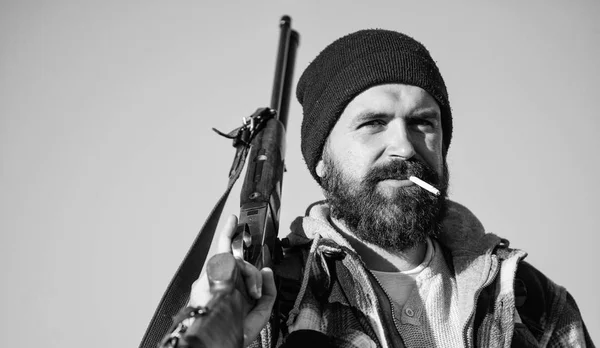 Hunting masculine hobby concept. Man brutal bearded guy gamekeeper blue sky background. Brutality and masculinity. Hunter with rifle gun close up. Guy bearded hunter spend leisure hunting and smoking