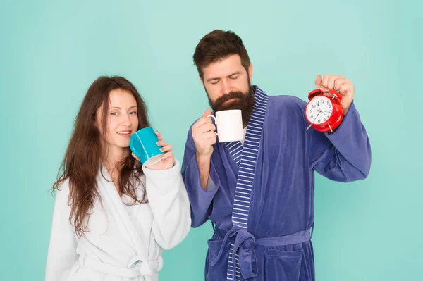 Homme barbu et femme endormie dégustent du café ou du thé le matin. C'est l'heure de se réveiller et de passer une bonne journée. Guy mari tient café et horloge. Chaque matin commence par un café. Couple en peignoirs avec tasses — Photo