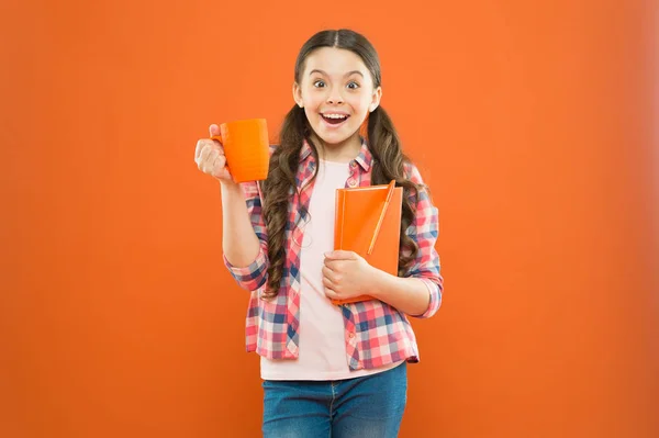 Με καύσιμα για το σχολείο. Μικρό κορίτσι απολαμβάνει το σχολείο διάλειμμά της σε πορτοκαλί φόντο. Ευτυχισμένο μαθήτρια που έχει σχολικό πρωινό το πρωί. Χαριτωμένο σχολείο παιδί χαμογελά με φλιτζάνι και βιβλίο σημειώσεων — Φωτογραφία Αρχείου