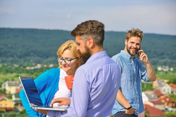 Colleagues with laptop work outdoor sunny day, nature background. Colleagues looking at screen laptop while man talking phone. Business partners meeting non formal atmosphere. Mobile internet concept