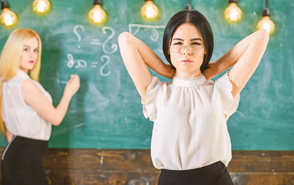 Mulheres atraentes preparando-se para a aula. Conceito de alunos e estagiários. Menina parece atraente enquanto senhora escrevendo no fundo quadro, desfocado. Estudante, professor de óculos em sala de aula — Fotografia de Stock