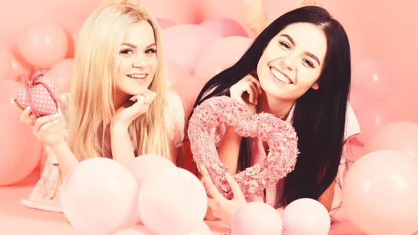 День святого Валентина концепция. Девочки лежали возле воздушных шаров, держали сердечные игрушки, розовый фон. Блондинка и брюнетка на улыбающихся лицах мечтают о любви и свидании. Сестры, друзья в пижаме на пижаме — стоковое фото