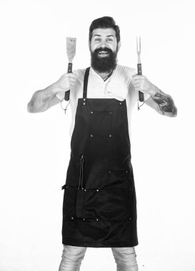 He is a great cook. Happy grill cook with cooking utensils. Bearded man holding fork and spatula for cooking and serving barbecue. Master cook wearing grilling apron. Chief cook in workwear. Cookout clipart