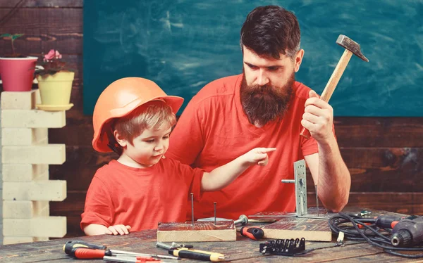 Niño, niño ocupado en casco protector aprendiendo a usar martillo con papá. Padre con barba enseñando a su hijo a usar herramientas, martilleo, pizarra de fondo. Concepto de paternidad — Foto de Stock