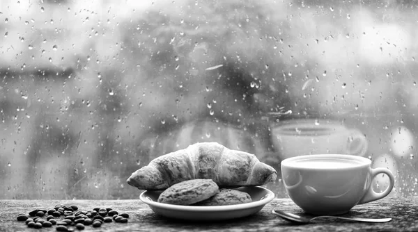 Koffie tijd op regenachtige dag. Vers gebrouwen koffie in witte kopje of mok op de vensterbank. Natte glazen raam en kopje warme cafeïne drank. Koffie drinken met croissant dessert. U geniet van koffie op regenachtige dag — Stockfoto
