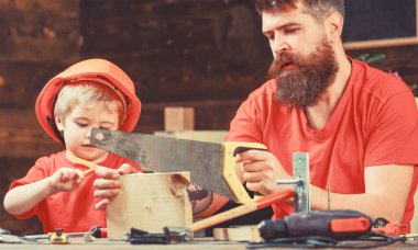 Çocuk, çocuk ile baba el testeresi kullanmayı öğrenme koruyucu kask içinde meşgul. Erkeksi görevleri kavramı. Baba, sakallı keskin el testeresi ile marangoz el sanatları testere için küçük oğlu öğretim üst