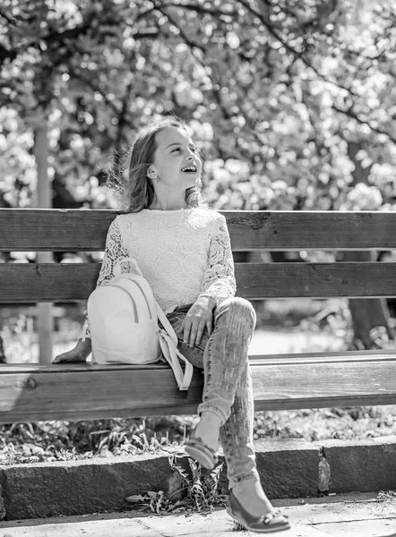 Dziewczyna na uśmiechniętą twarz siedzi na ławce, drzewa sakura na rozmytym tłem. Relaks podczas spaceru w parku, w pobliżu wiśniowy kwiat dziewczyna. Wiosna idzie koncepcja. Słodkie dziecko z plecaka cieszyć się słoneczny wiosenny dzień — Zdjęcie stockowe