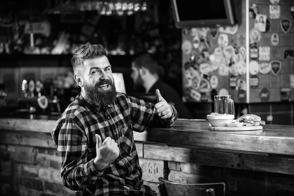 High calorie snack. Hipster relaxing at pub. Pub is relaxing place to have drink and relax. Brutal hipster bearded man sit at bar counter. Man with beard drink beer eat burger menu. Enjoy meal in pub