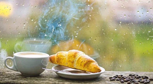 Coffee drink with croissant dessert. Enjoying coffee on rainy day. Coffee time on rainy day. Fresh brewed coffee in white cup or mug on windowsill. Wet glass window and cup of hot caffeine beverage