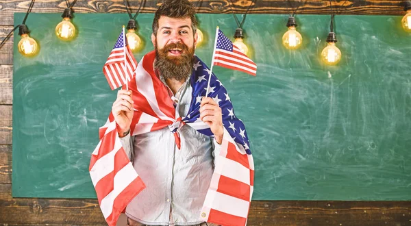 Conceito de educação patriótica. Homem com barba e bigode no rosto feliz segura bandeiras dos EUA, em sala de aula, quadro-negro no fundo. Professora americana acena com bandeiras americanas. Programa de intercâmbio de estudantes — Fotografia de Stock