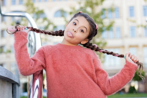 A divertir-se com o cabelo dela. Menina engraçada segurando tranças de cabelo no fundo urbano. Menina bonito pequeno com longo cabelo morena mostrando língua ao ar livre. Modelo de cabelo pequeno com olhar louco — Fotografia de Stock