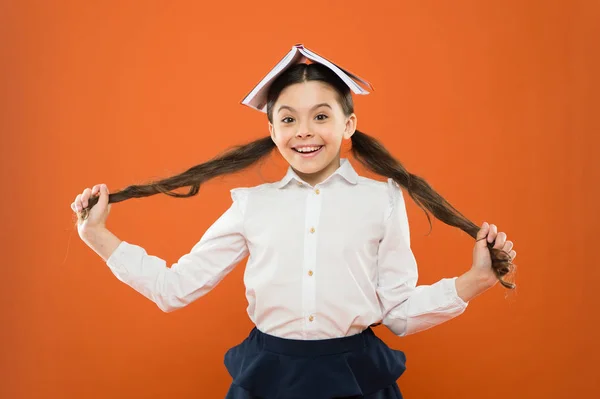 Let your hair speak for itself. Cute little girl holding long brunette hair on orange background. Small school child with book on head wearing hair in ponytails. Hair styling and haircare for school — Stock Photo, Image