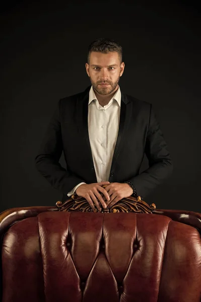 Businessman or man in formal suit on dark background. Man on serious face posing behind leather armchair. Business success concept. Man with bristle looks confident and successful