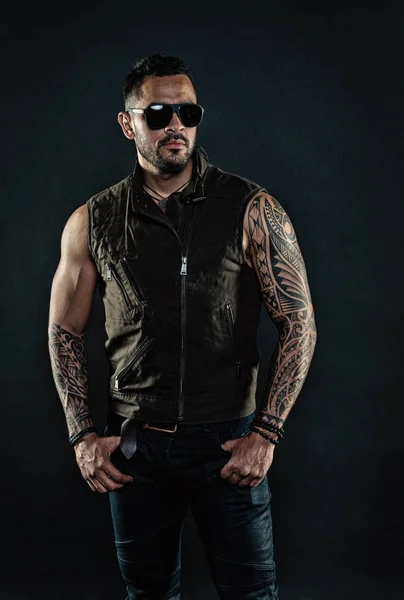 Fashion macho in trendy sunglasses. Tattoo model with beard on unshaven face. Bearded man with tattoo on strong arms. Tattooed man with biceps and triceps. Fashion style and trend, vintage filter