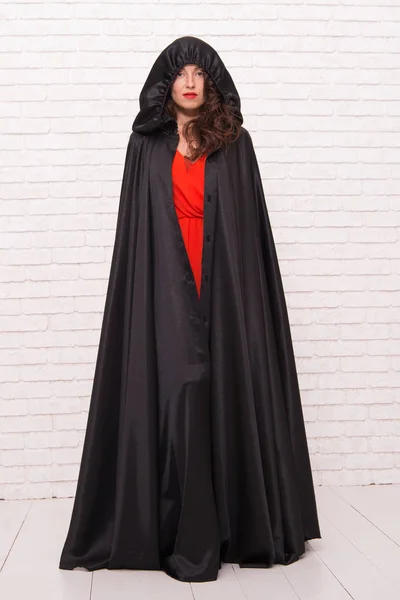 Scary but iconic. A woman witch. Sexy woman wearing halloween costume on white brickwall. Sensual woman with classic witch look. Mysterious woman in hooded black cloak