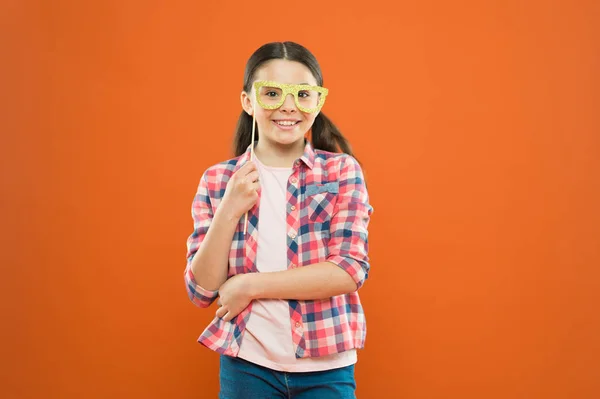 Περίεργο βλέμμα. Αξιολάτρευτο κορίτσι χαμογελά με το κόμμα βλέμμα σε πορτοκαλί φόντο. Χαριτωμένο μικρό παιδί που έχει χαρούμενο βλέμμα μέσα από τα γυαλιά. Μόδα εμφάνιση του μικρού μοντέλου μόδας — Φωτογραφία Αρχείου