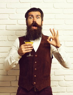 Uzun sakallı ve bıyıklı yakışıklı bir adam. Şaşırmış yüzünde şık saçlar var. Elinde klasik süet deri yelek, beyaz tuğla duvar arka planında alkollü içecek bardağı var.
